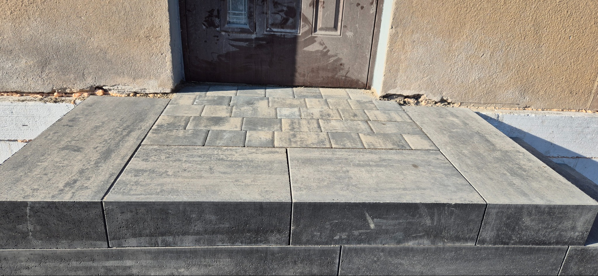 Schody wejściowe wyłożone kostką brukową w odcieniach szarości, prowadzące do ciemnych drzwi. Widoczne fragmenty ściany budynku z różną fakturą tynku.