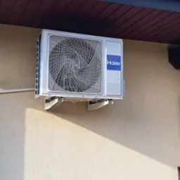 Zewnętrzna jednostka klimatyzatora Haier zamontowana na elewacji domu z widocznym fragmentem drewnianej pergoli i rurą spustową.