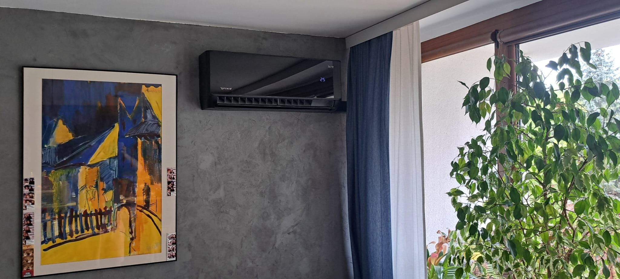 Czarny klimatyzator Rotenso zamontowany na szarej, strukturalnej ścianie w salonie, obok okna z zasłonami i rośliną doniczkową, pod nim abstrakcyjny obraz w ramie.