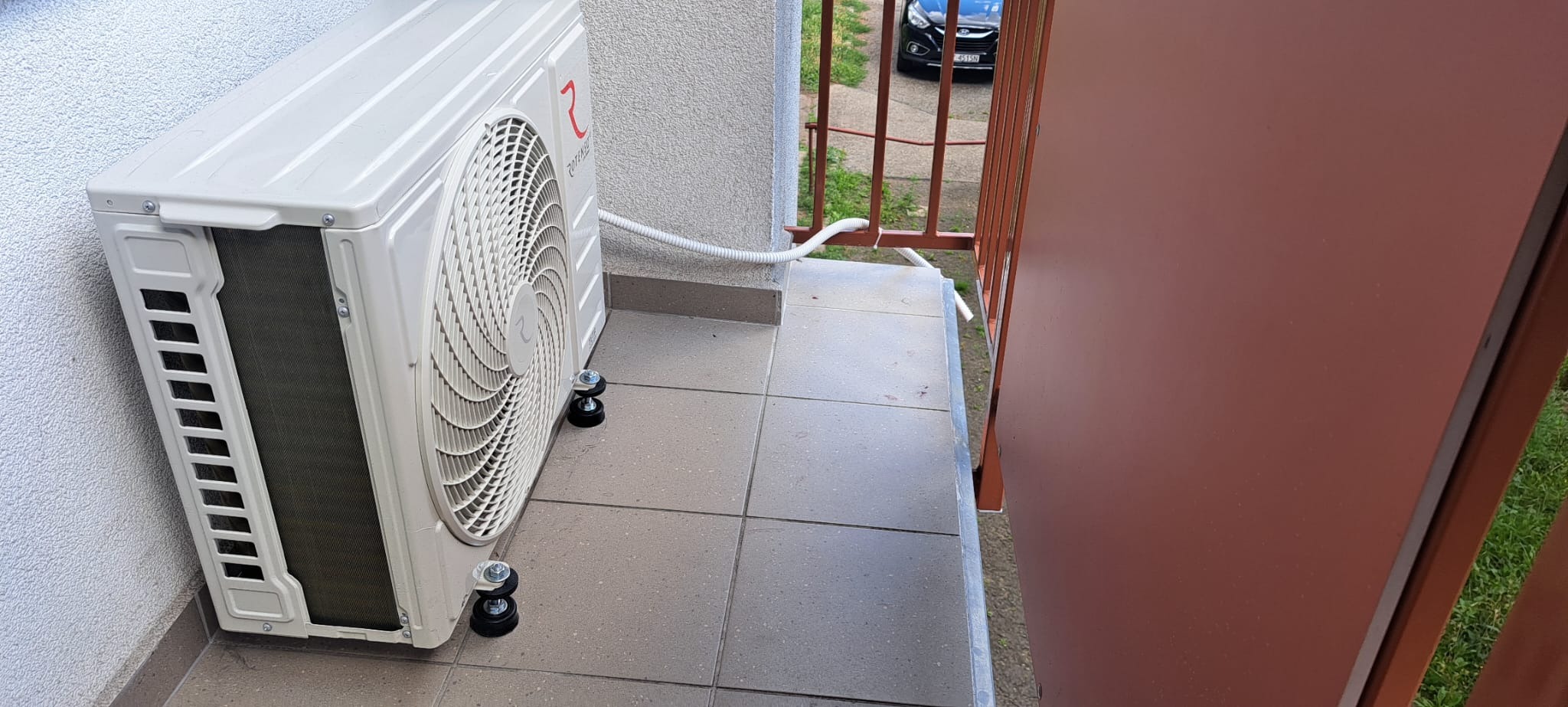 Biała jednostka zewnętrzna klimatyzatora, umieszczona na balkonie na specjalnych podkładkach antywibracyjnych, z widocznym przewodem odprowadzającym skropliny.