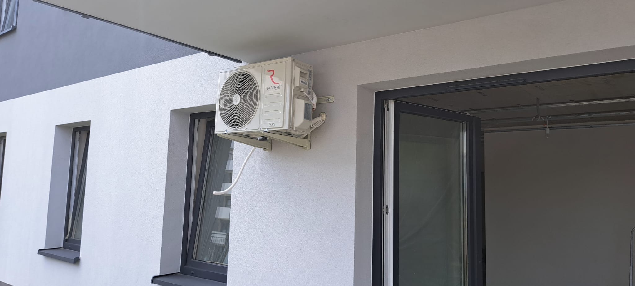 Biała jednostka zewnętrzna klimatyzatora Rotenso zamontowana na ścianie budynku z widocznymi oknami i otwartym balkonem z surowym wnętrzem.