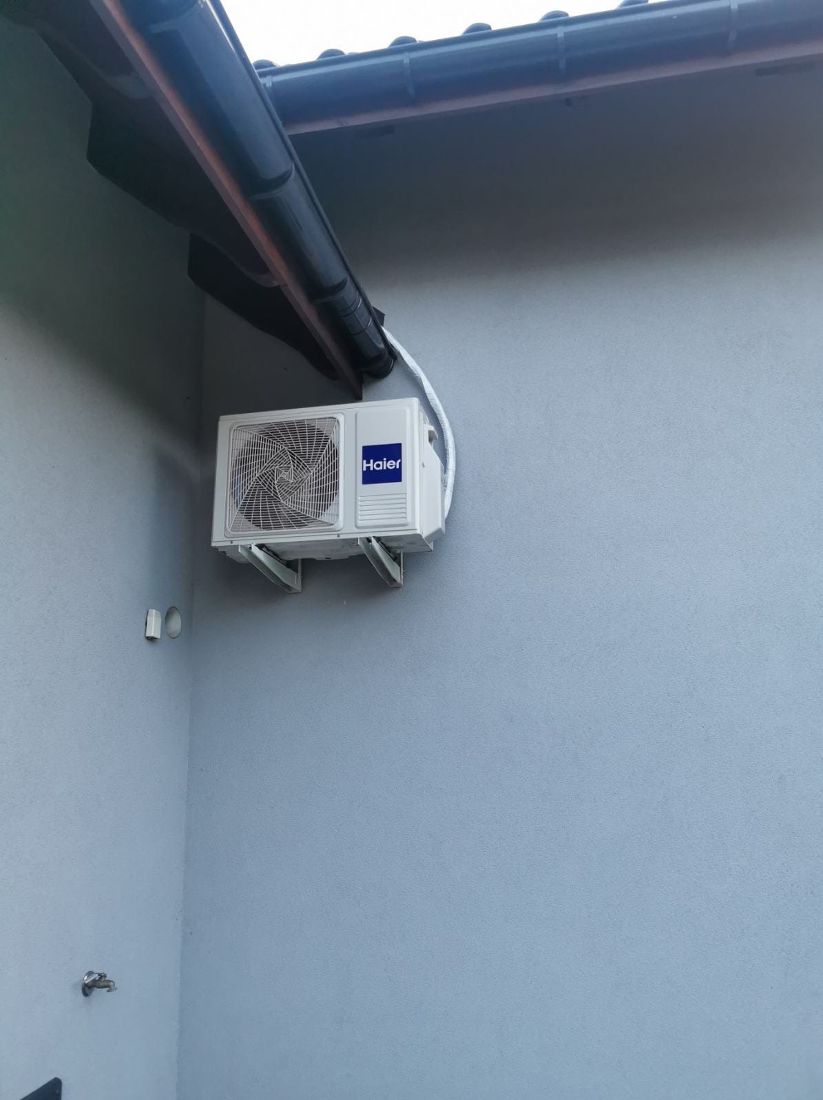 Biała jednostka zewnętrzna klimatyzatora Haier zamontowana na szarej ścianie budynku pod rynną dachową.