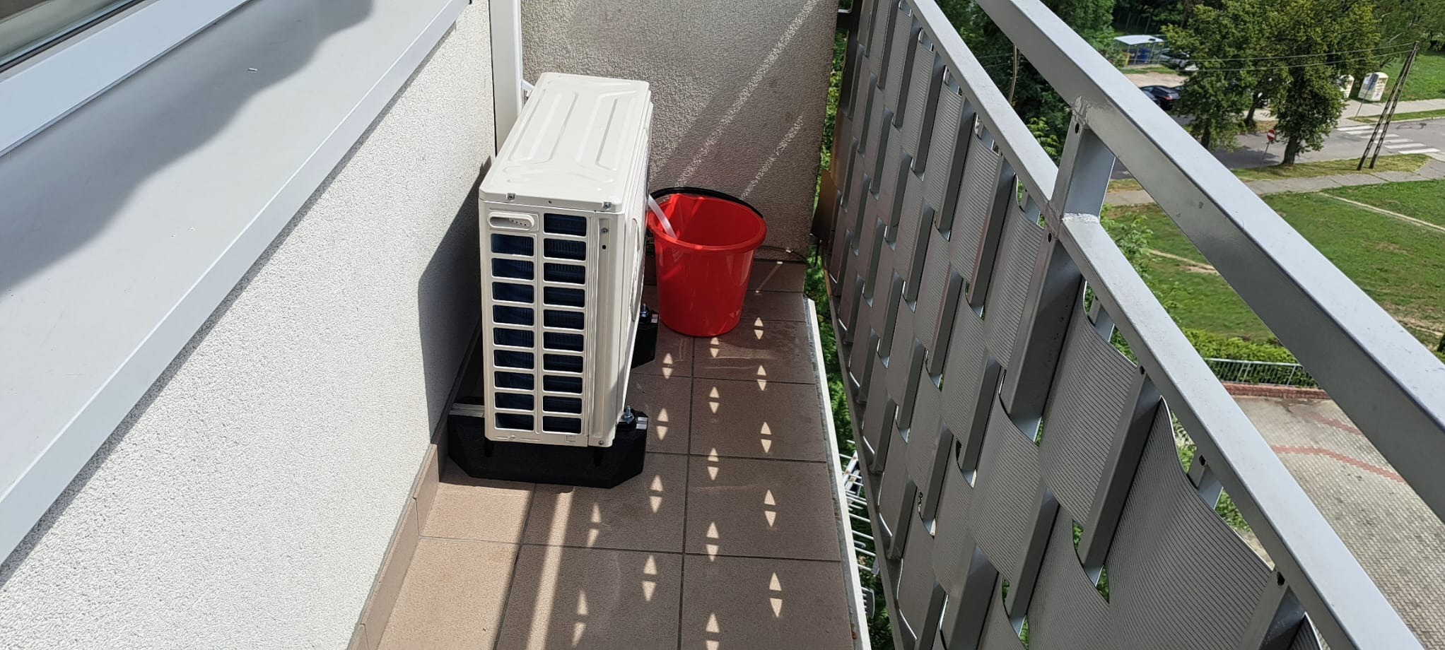 Biała jednostka zewnętrzna klimatyzatora na balkonie z terakotą, obok czerwone wiadro, widok na zieleń za balustradą.