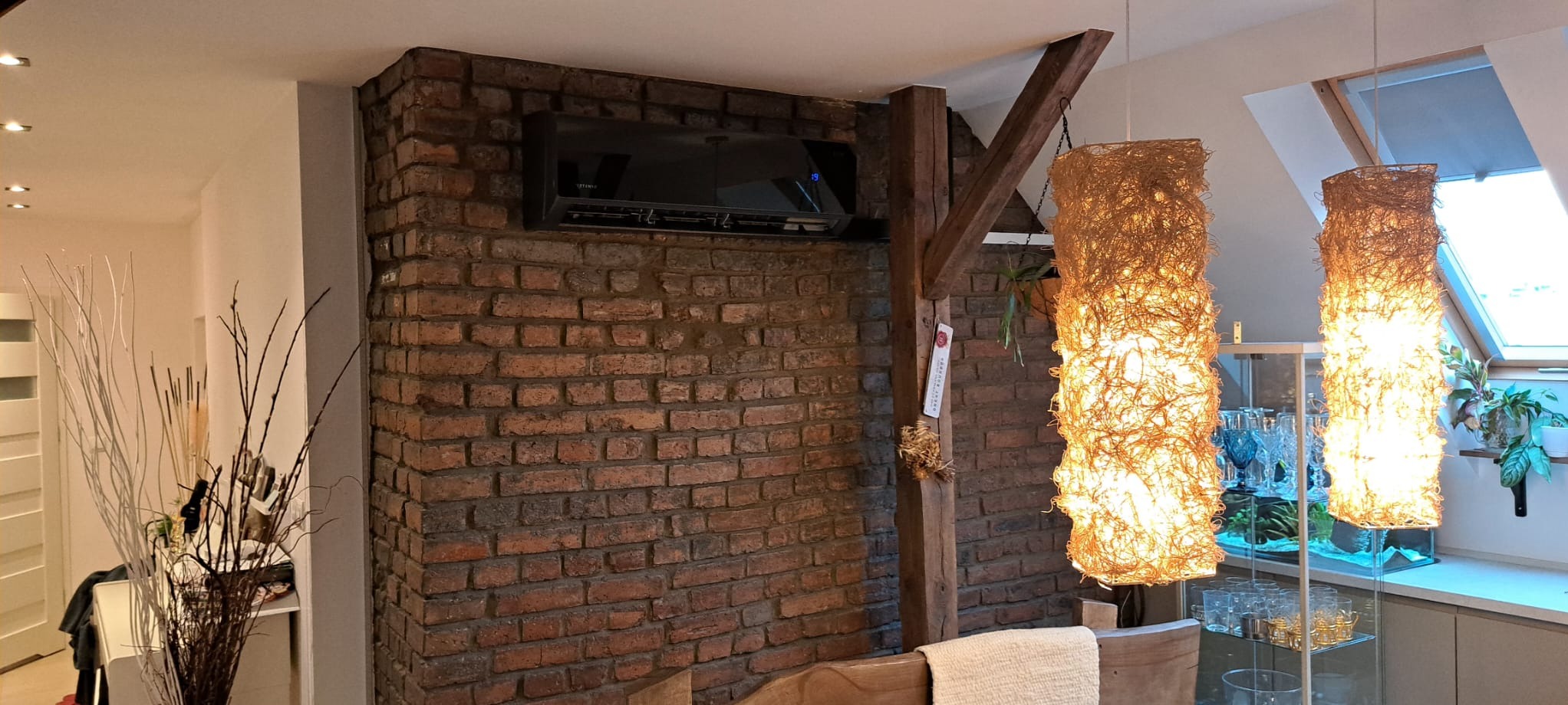 Czarny klimatyzator ścienny zamontowany na ceglanej ścianie w nowoczesnym wnętrzu z drewnianymi belkami i dekoracyjnymi lampami.