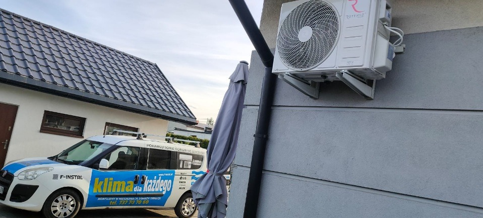 Biała jednostka zewnętrzna klimatyzacji Rotenso zamontowana na szarej ścianie domu, widoczny samochód firmowy z reklamą klimatyzacji i parasol ogrodowy.