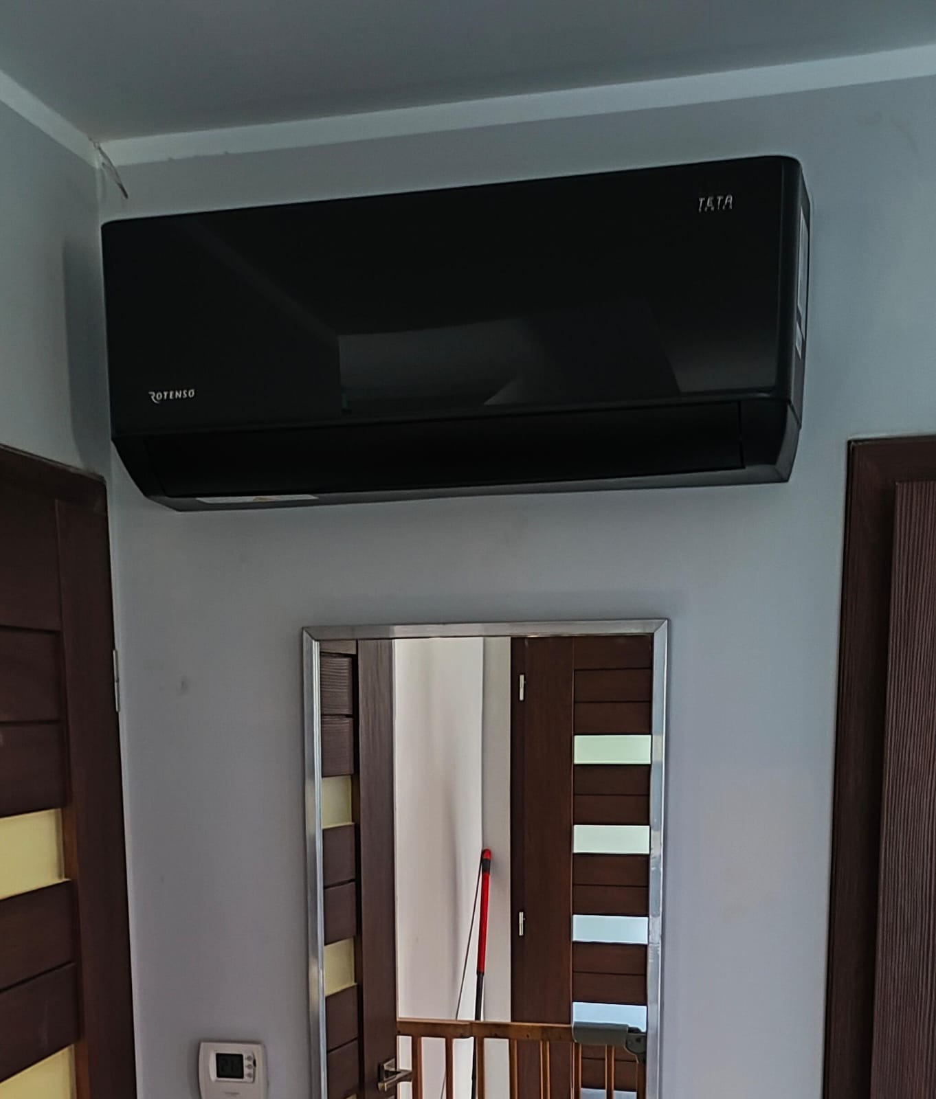 Czarny klimatyzator marki Rotenso TATA zamontowany na szarej ścianie, widoczne drzwi po bokach i lustro z odbiciem wnętrza.