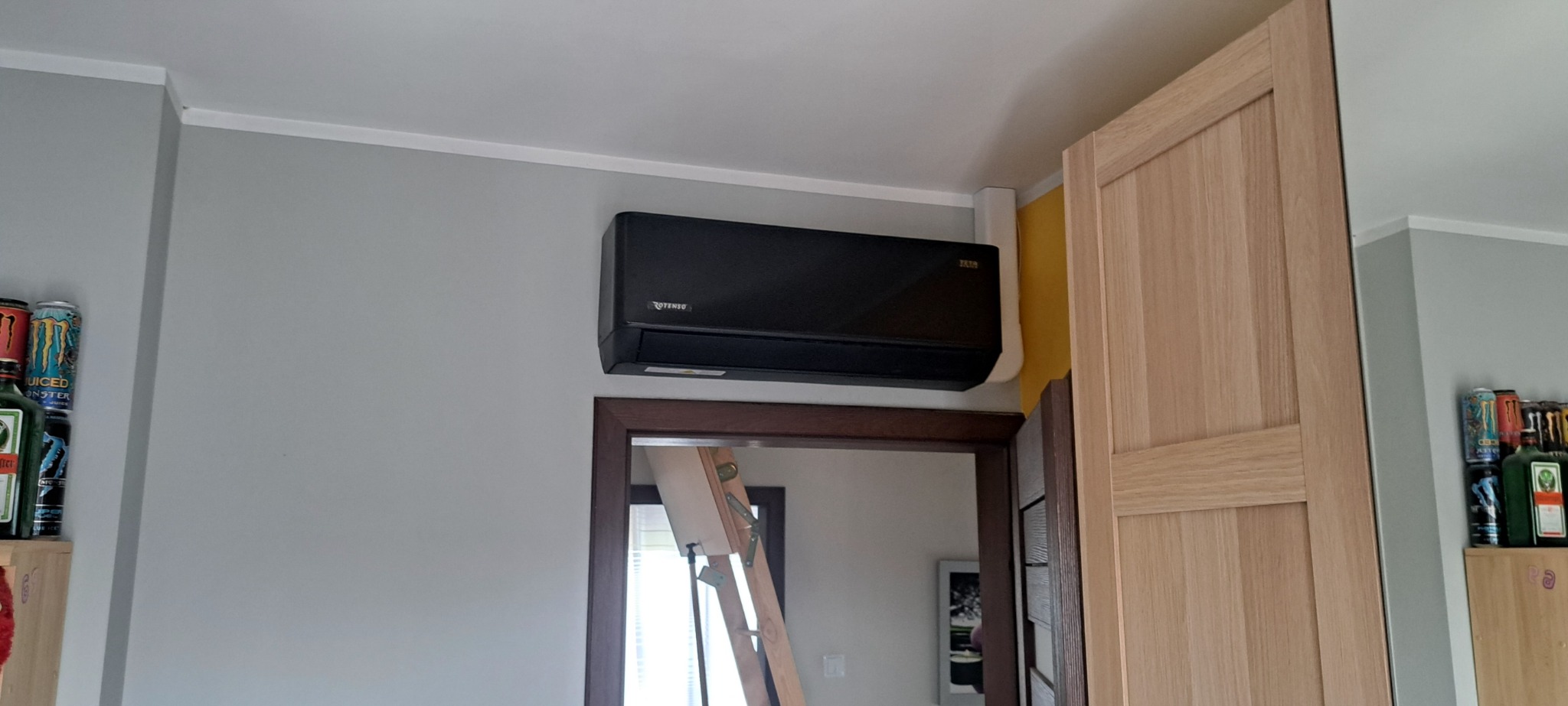 Czarny klimatyzator Rotenso zamontowany nad drzwiami, widoczna drabina w odbiciu lustra i regał z napojami energetycznymi po lewej stronie.
