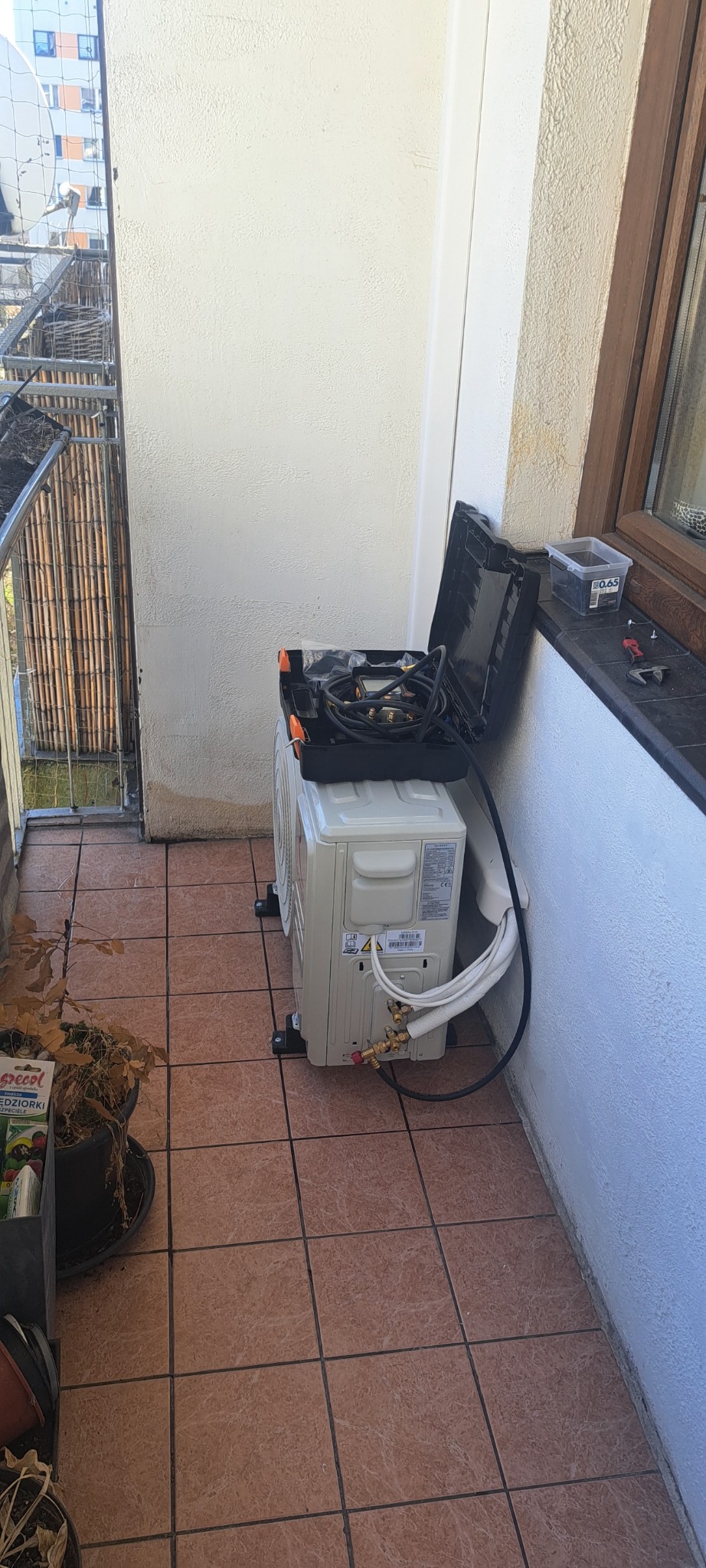 Jednostka zewnętrzna klimatyzacji na balkonie, obok skrzynka z narzędziami, widoczne rurki i kable podłączeniowe.