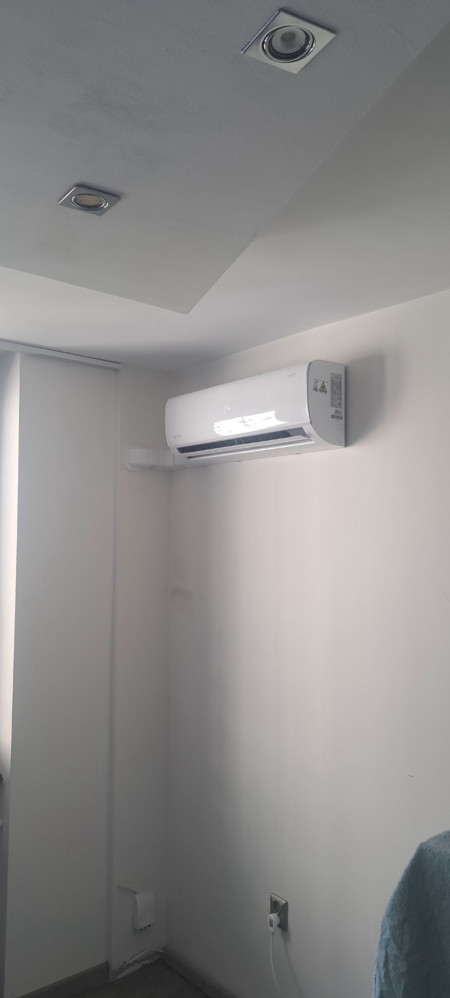 Biały klimatyzator ścienny z wyświetlaczem temperatury (24 stopnie) zamontowany w rogu pomieszczenia z białymi ścianami, widoczne gniazdko elektryczne i lampy sufitowe.