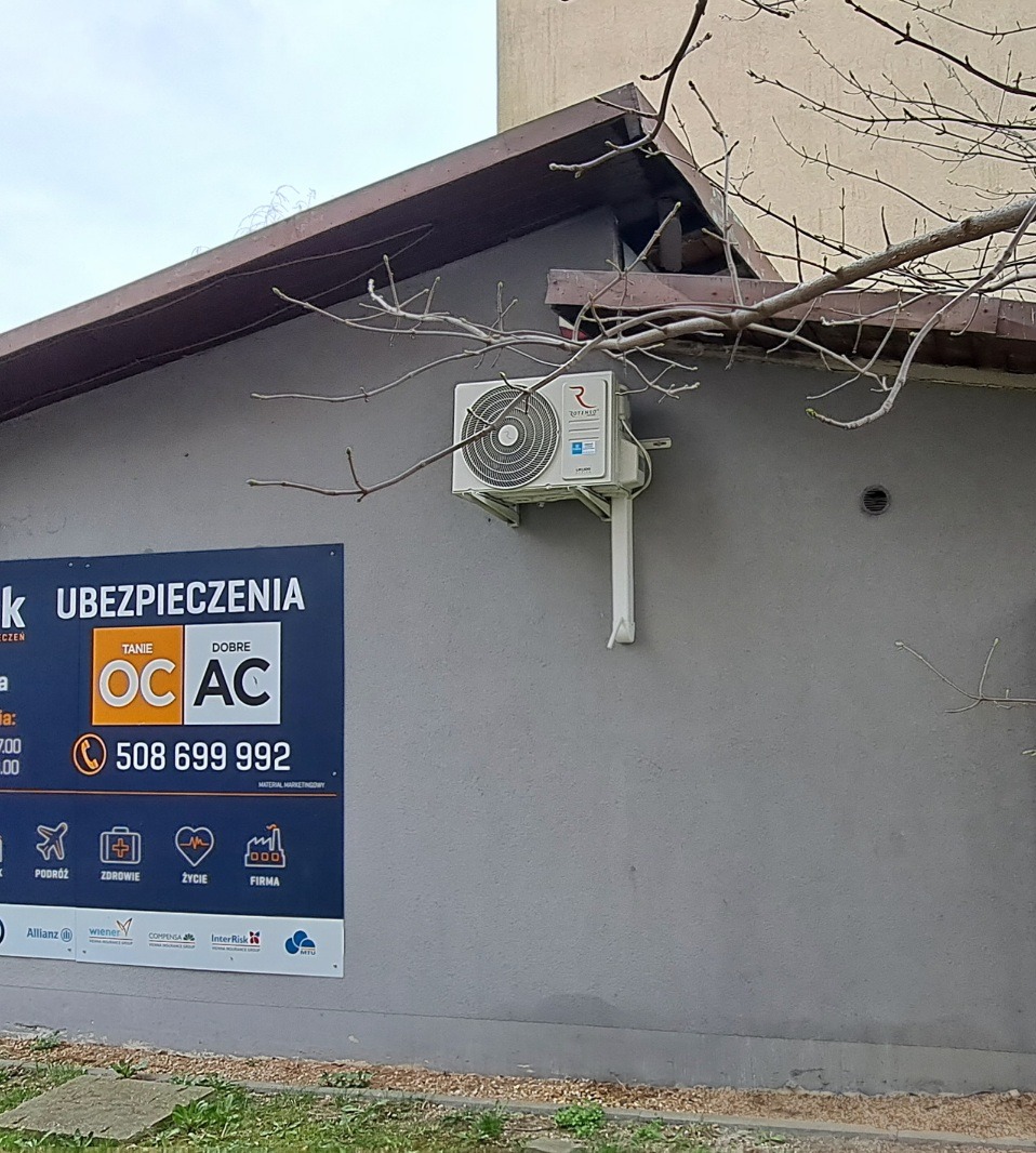 Klimatyzator ROTENSO zamontowany na szarej ścianie budynku w Bytomiu, widoczny fragment reklamy ubezpieczeń oraz gałęzie drzewa.