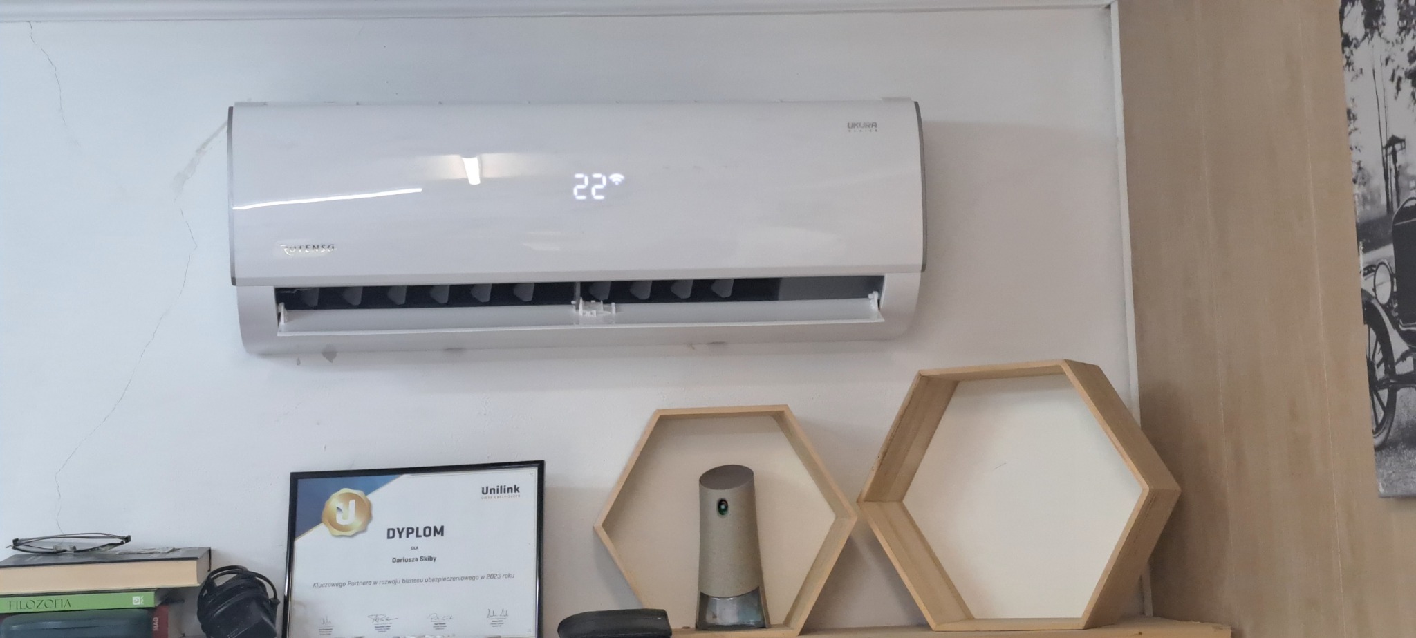 Biały klimatyzator ścienny marki Tensg Ukura z wyświetlaną temperaturą 22 stopnie Celsjusza, zamontowany w pomieszczeniu z dekoracyjnymi półkami w kształcie plastra miodu i dyplomem na ścianie.