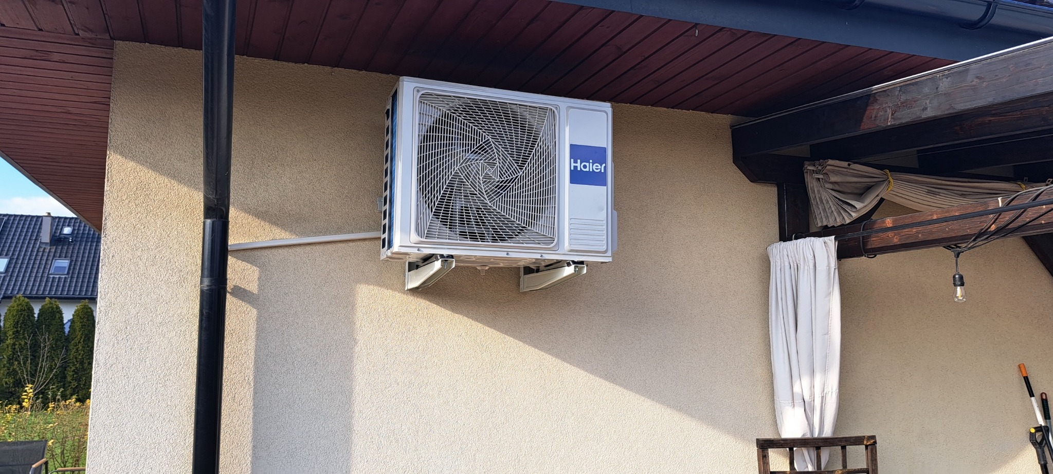 Zewnętrzna jednostka klimatyzatora Haier zamontowana na elewacji domu z widocznym fragmentem drewnianej pergoli i rurą spustową.