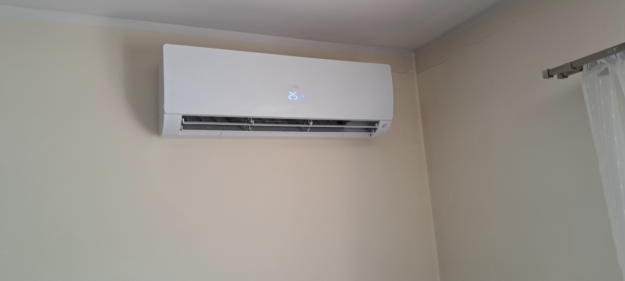 Nowoczesny, biały klimatyzator ścienny z wyświetlaczem temperatury 25 stopni Celsjusza, zamontowany w rogu pokoju na beżowej ścianie z widocznym pęknięciem przy suficie.
