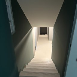 VS Group - Widok z góry na świeżo pomalowane schody prowadzące do korytarza z jasną podłogą, ściany w odcieniach szarości i zieleni, zabezpieczone folią schody na dole.