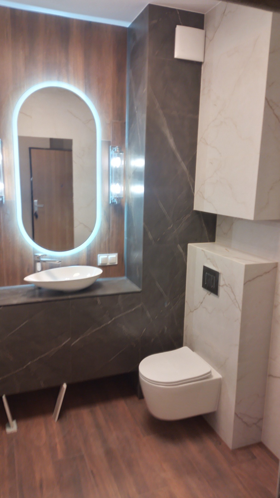 Nowoczesna łazienka z podwieszaną toaletą, umywalką nablatową i podświetlanym lustrem, wykończona płytkami imitującymi marmur i drewno.