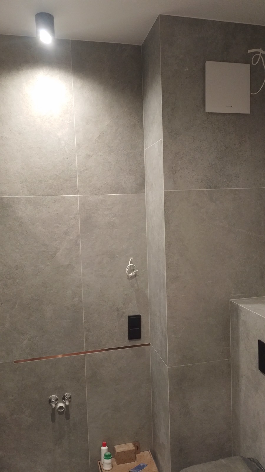 Wykończona łazienka z szarymi płytkami imitującymi kamień, widoczne przyłącza wodne, akcesoria oraz fragment podestu z materiałami budowlanymi.