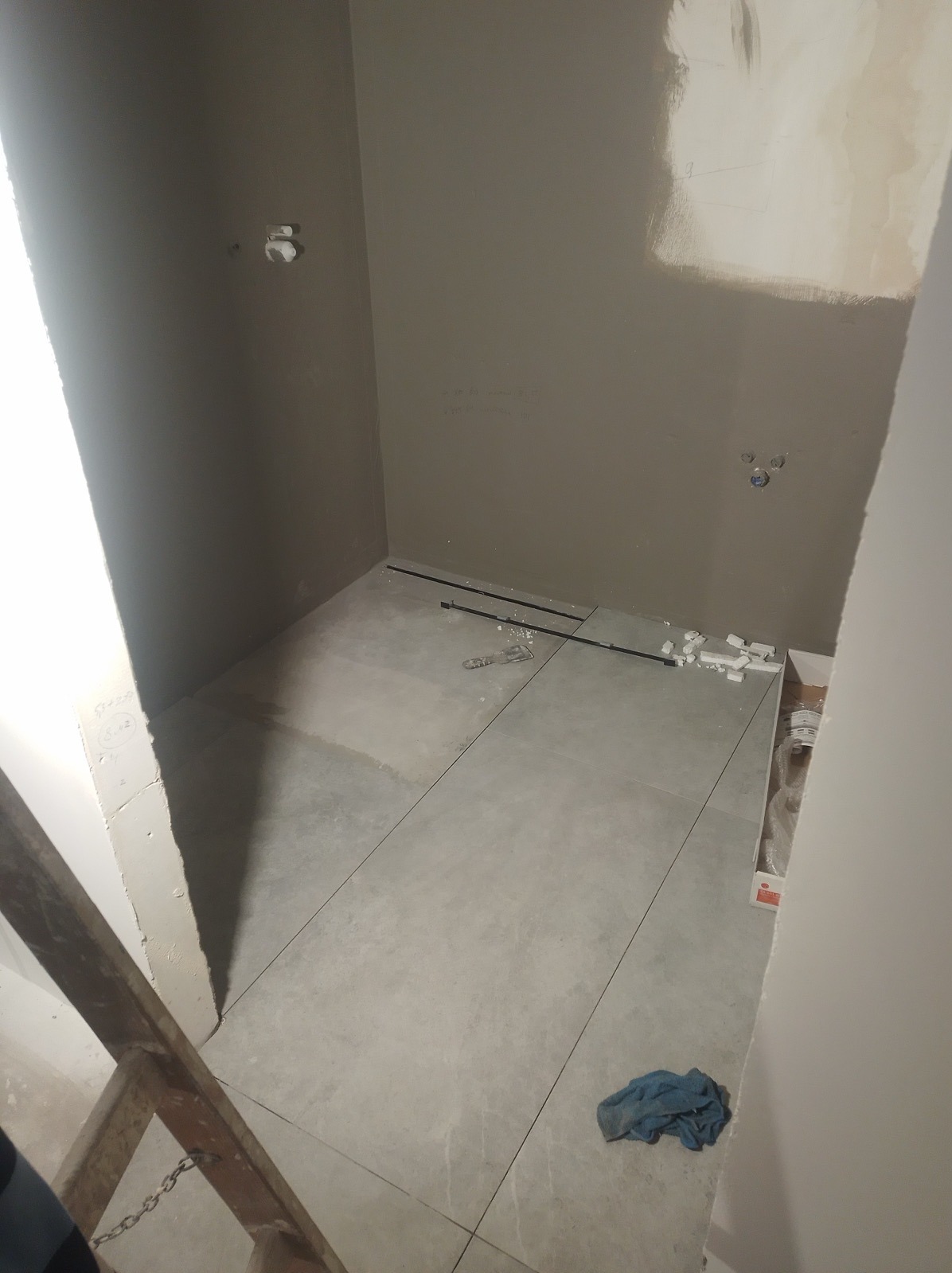 Wnętrze łazienki w trakcie układania szarych płytek podłogowych, widoczne listwy progowe, resztki zaprawy i narzędzia, ściany przygotowane do malowania, fragment drabiny malarskiej.