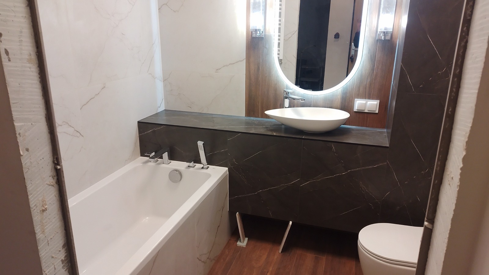 Nowoczesna łazienka w trakcie wykańczania: biała wanna, umywalka nablatowa na blacie z ciemnego marmuru, okrągłe lustro z podświetleniem LED, widoczne niedokończone wykończenie ściany.
