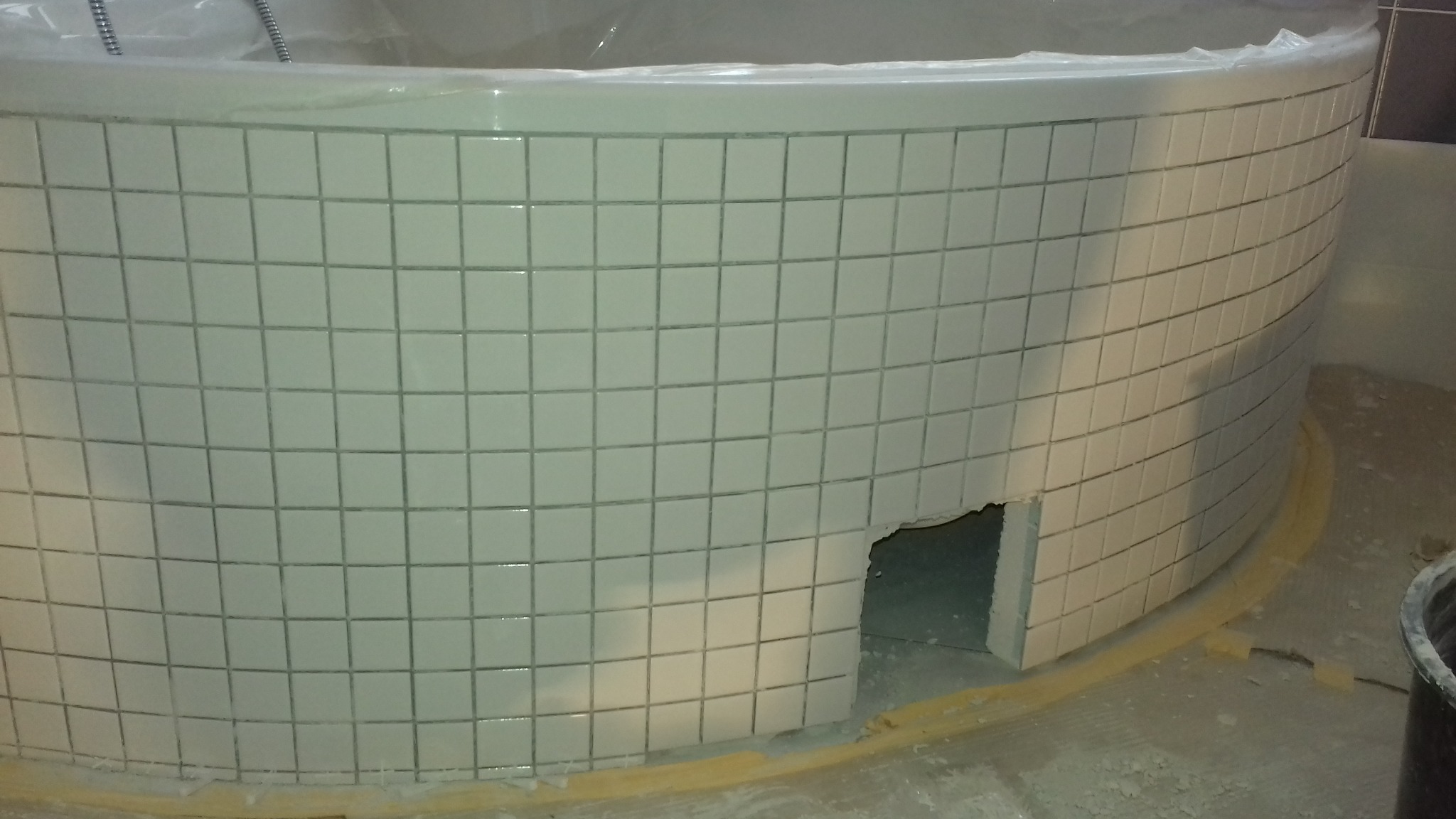 Okrągła wanna w trakcie obkładania jasnymi, kwadratowymi płytkami; widoczny otwór rewizyjny w dolnej części obudowy wanny i zabezpieczona folią górna krawędź wanny.