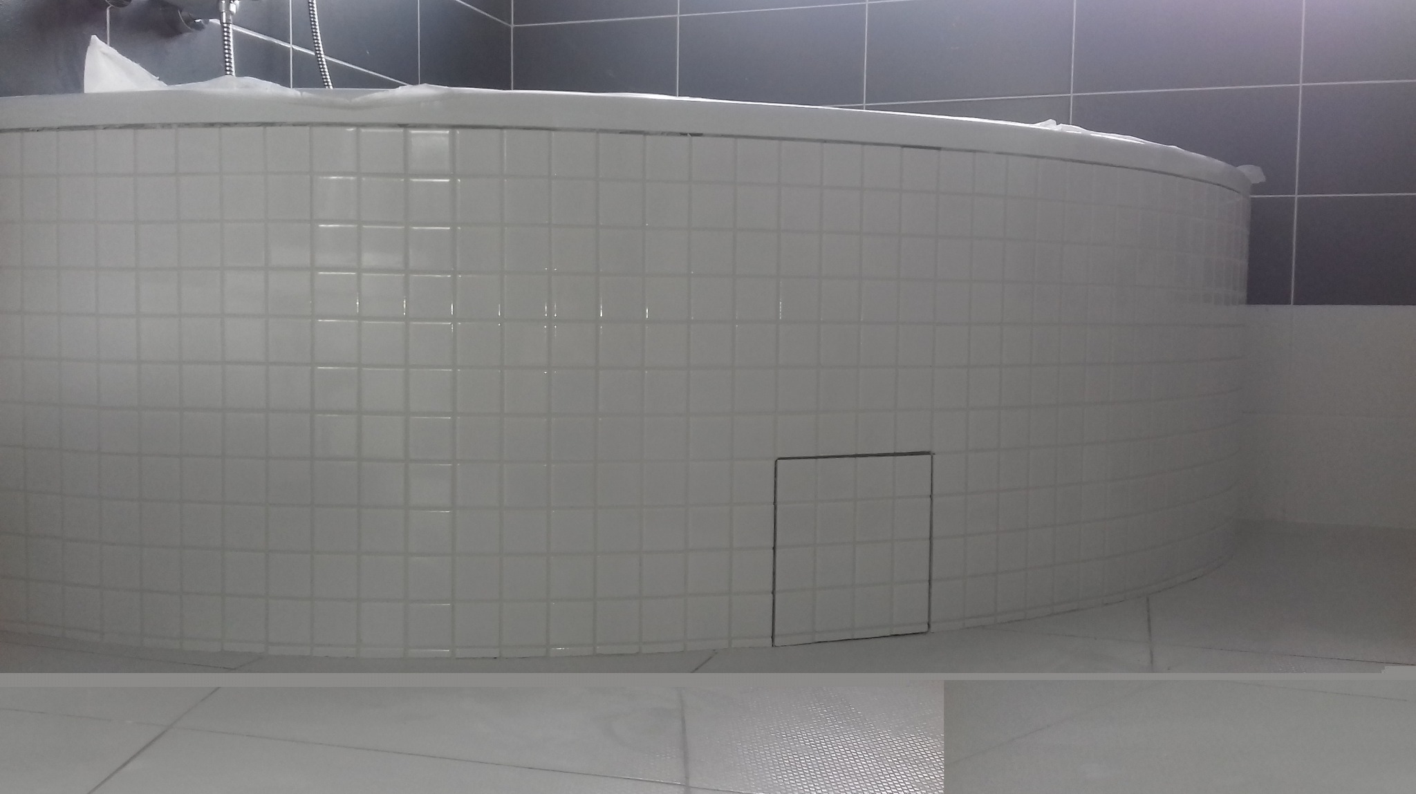 Okrągła wanna obłożona białymi, kwadratowymi płytkami z małą klapką rewizyjną, widoczna część szarej ściany z większymi płytkami w tle, zabezpieczona folią ochronną.