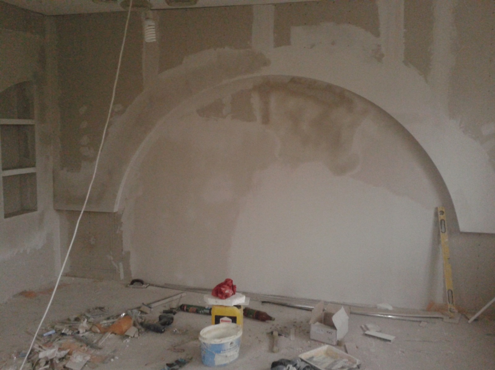 Ściana w trakcie szpachlowania z widocznym łukiem, narzędzia i materiały budowlane porozrzucane na podłodze, poziomica oparta o ścianę.