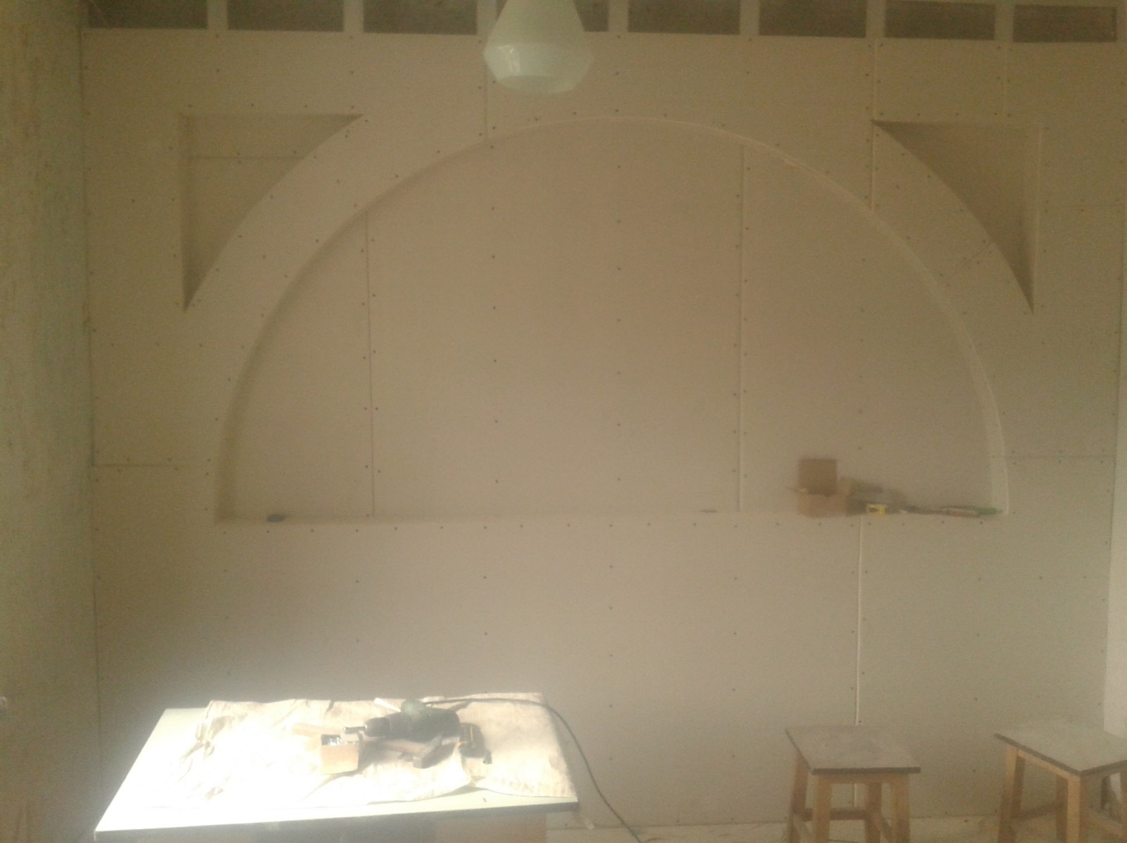 Ściana z płyt gipsowo-kartonowych w trakcie zabudowy, z wyciętymi półokrągłymi i trójkątnymi niszami, widoczne narzędzia i stoliki robocze.