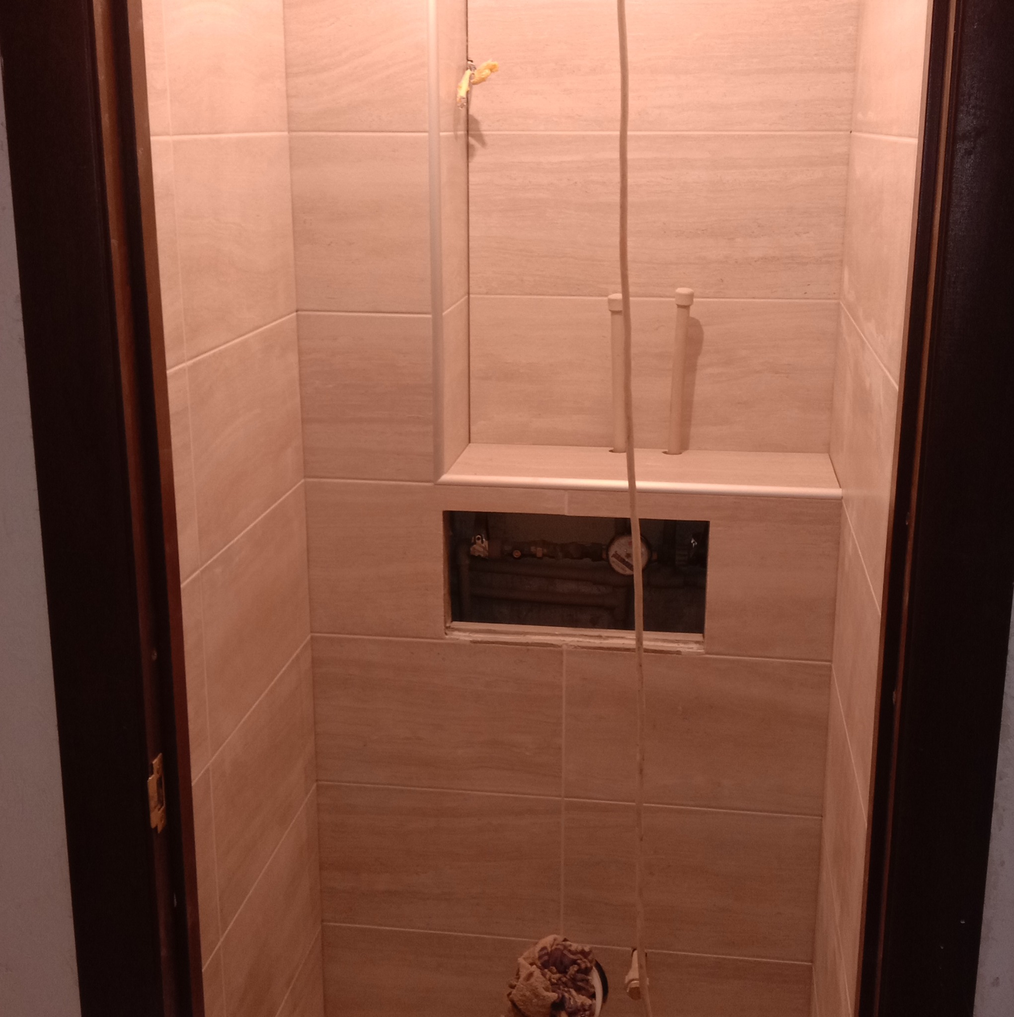 Wnęka z beżowymi płytkami w łazience, widoczne rury instalacji wodnej i licznik, niedokończone prace, widok zza otwartych drzwi.