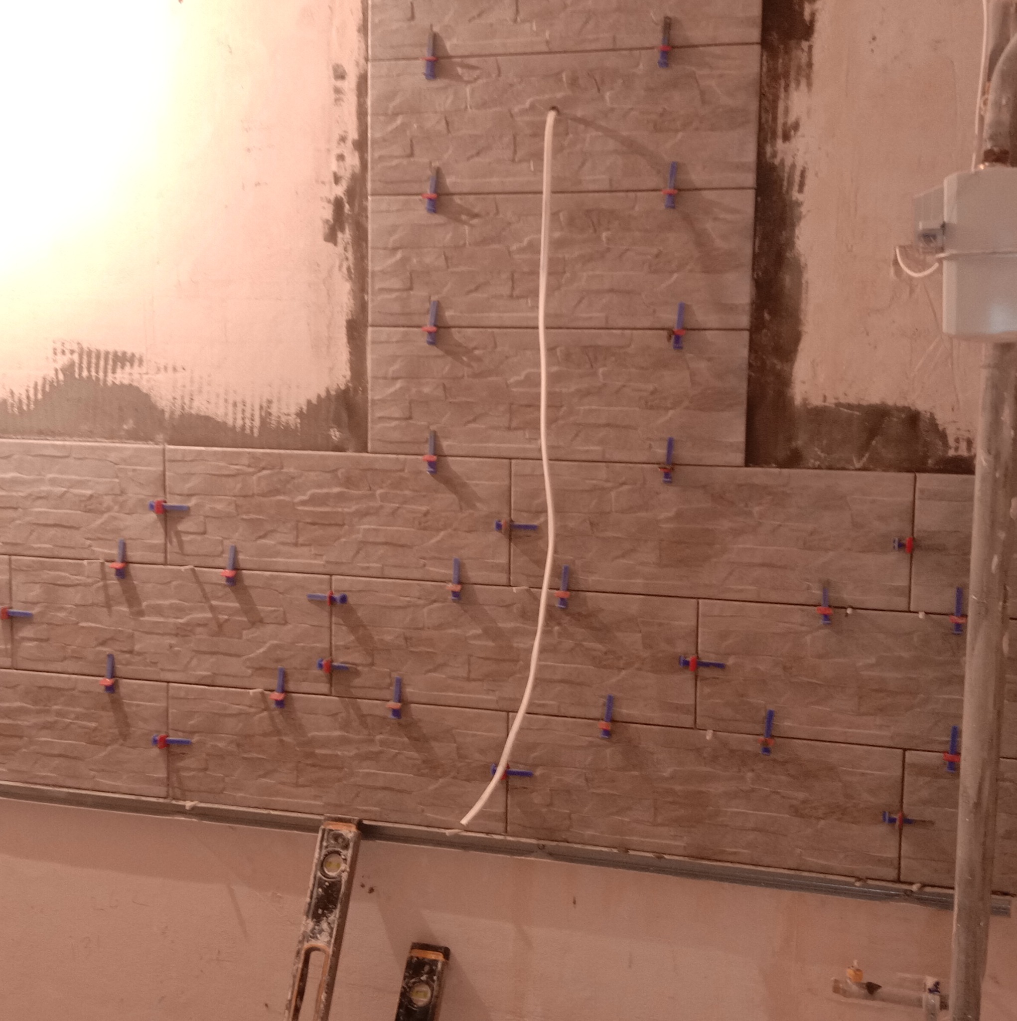 Ściana w trakcie układania płytek imitujących kamień, z użyciem klipsów poziomujących, widoczny kabel wystający ze ściany oraz poziomica przy ścianie.