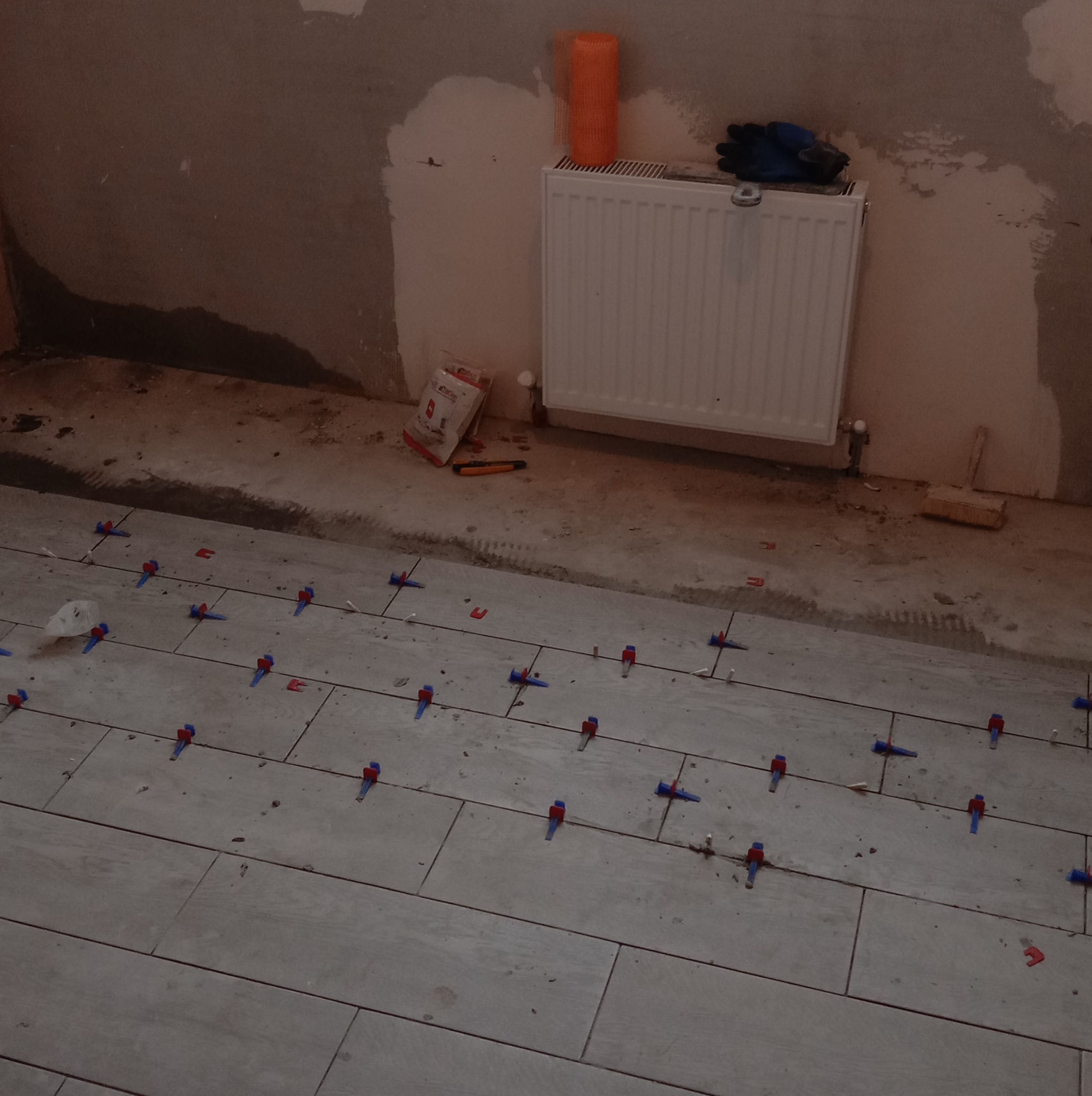 Układanie płytek podłogowych z użyciem systemu poziomowania w Płońsku, widoczne krzyżyki dystansowe, obok grzejnik i narzędzia glazurnicze.