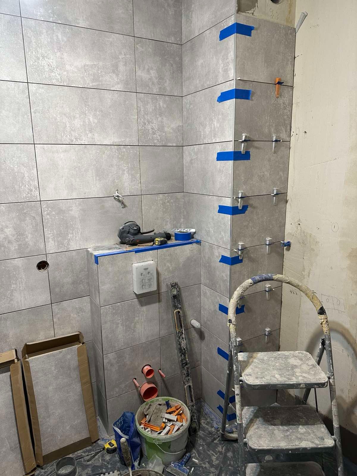 Ściany łazienki w trakcie układania szarych płytek, zabezpieczone niebieską taśmą i klipsami poziomującymi, widoczna drabina, poziomica i narzędzia glazurnicze.