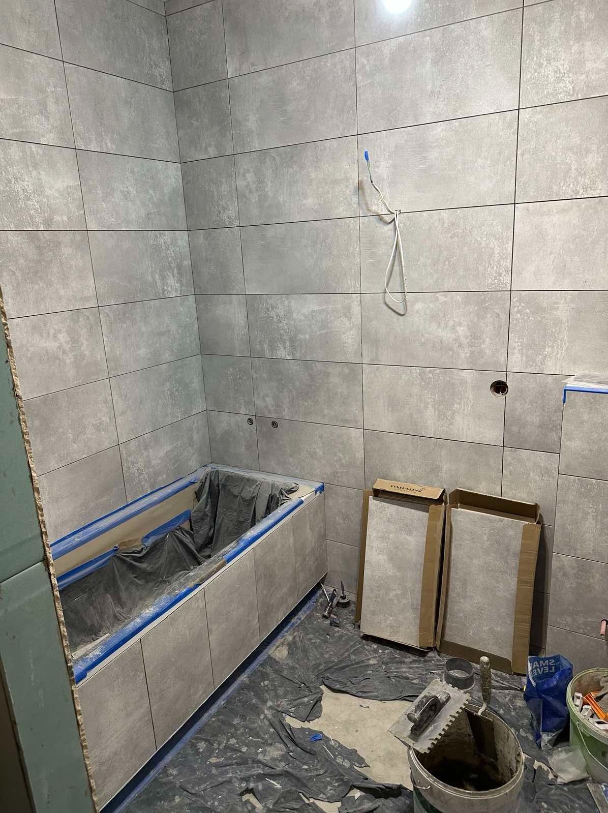 Łazienka w trakcie wykańczania: szare, prostokątne płytki imitujące beton na ścianach, obudowana wanna zabezpieczona folią i taśmą malarską, narzędzia glazurnicze na podłodze przy kartonach...