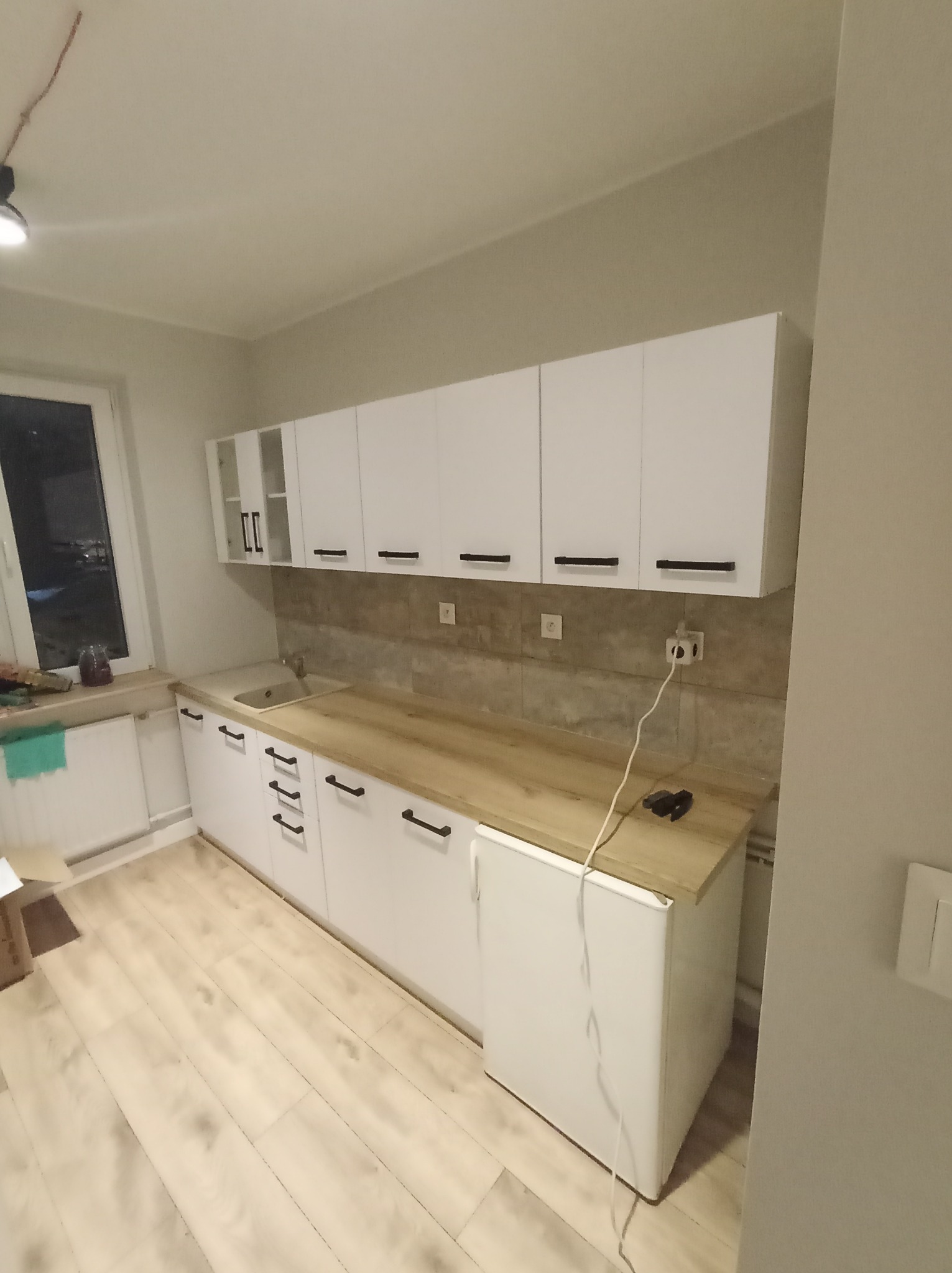 Nowoczesna, biała zabudowa kuchenna z drewnianym blatem, czarnymi uchwytami i beżowymi płytkami na ścianie, widok na okno i jasną podłogę.