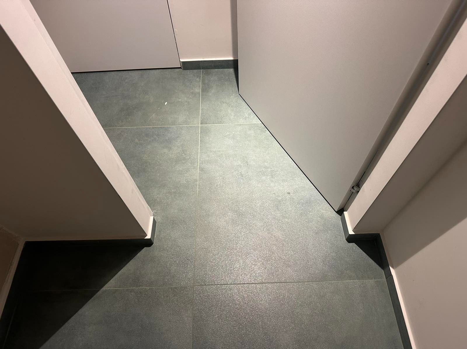 Szare płytki podłogowe ułożone w kwadraty, widoczne fragmenty białych ścian i ościeżnicy z zamontowanymi drzwiami.