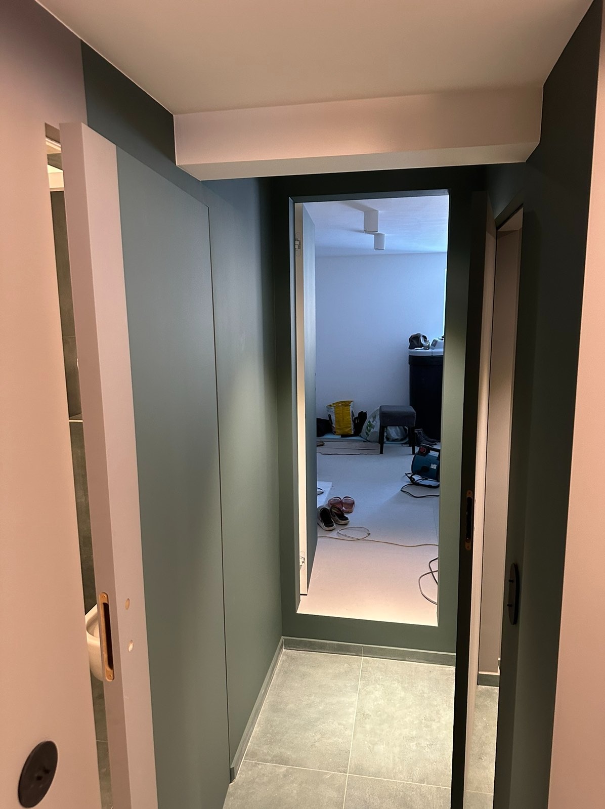 Wnętrze w trakcie prac wykończeniowych: widok przez otwarte drzwi na pokój z workiem, narzędziami i fragmentem podłogi z szarych płytek.