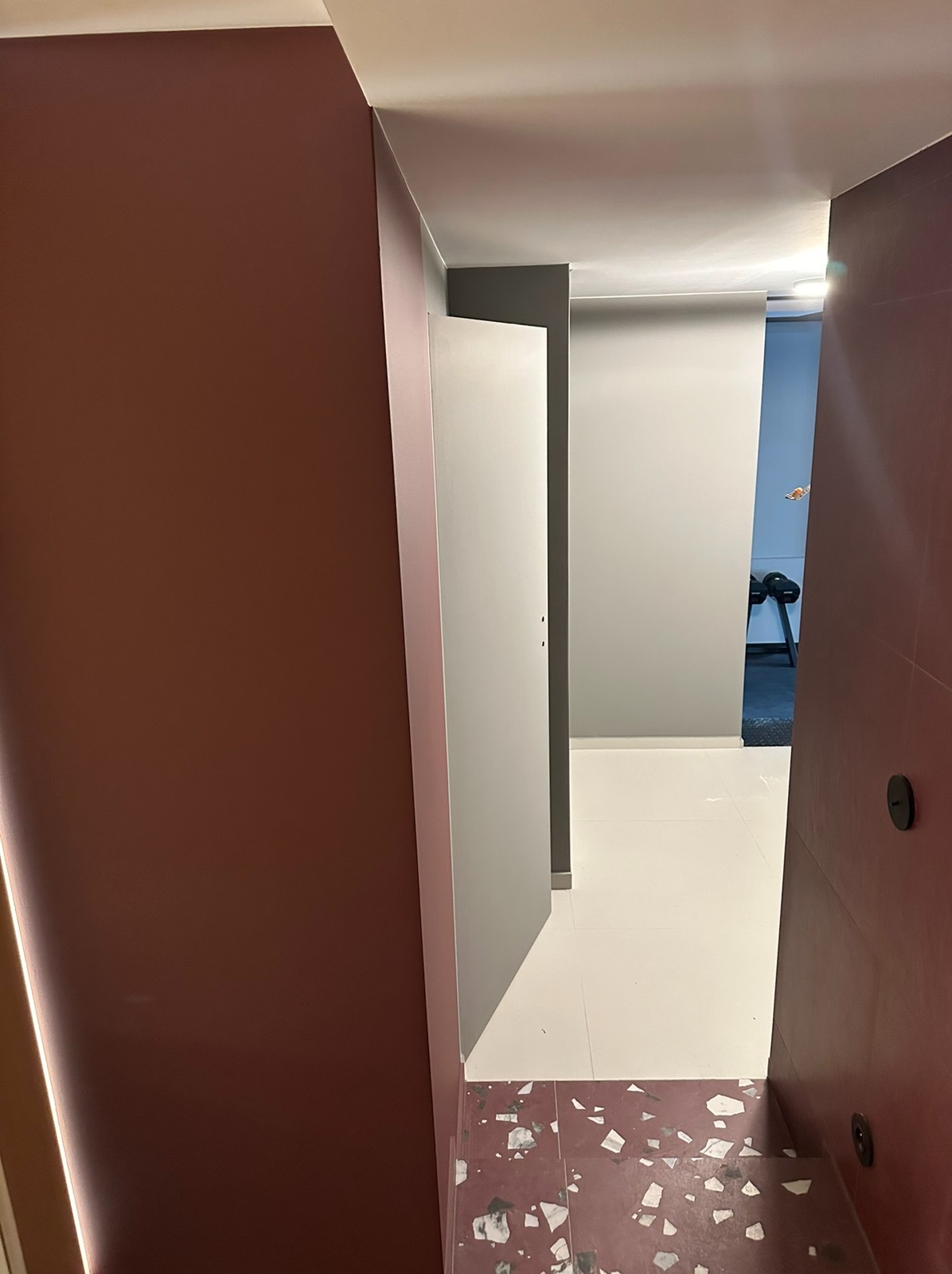 Korytarz po odświeżeniu z bordowymi ścianami, białymi płytkami podłogowymi i bordowymi schodami z mozaiką, widok na szare pomieszczenie z siłownią w tle.