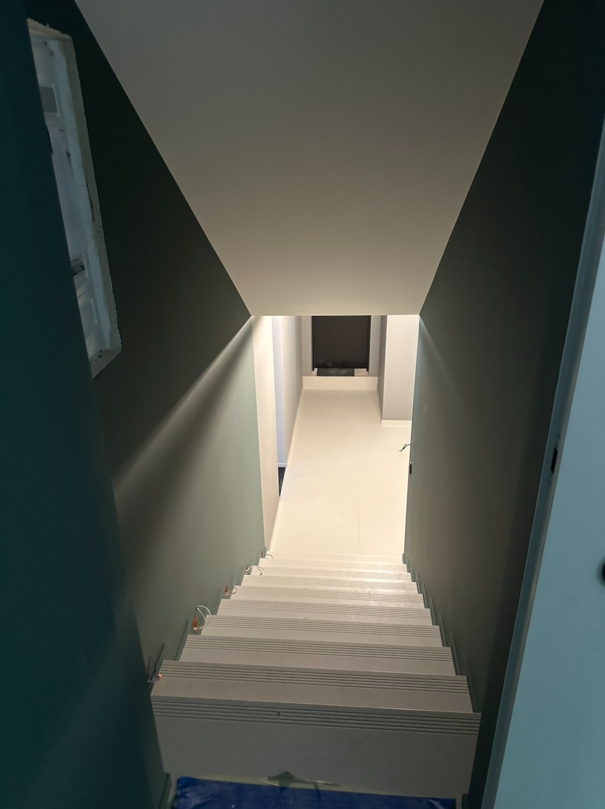Widok z góry na świeżo pomalowane schody prowadzące do korytarza z jasną podłogą, ściany w odcieniach szarości i zieleni, zabezpieczone folią schody na dole.