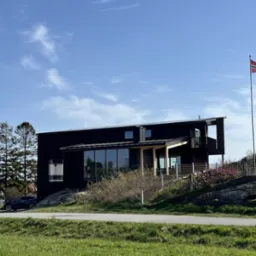 Nowoczesny dom z czarną elewacją, dużymi oknami i balkonem, otoczony zielenią i norweską flagą na maszcie.