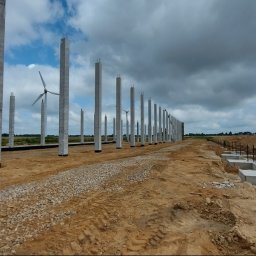 Biuro Inżynierskie Jakub Szewczyk - Wznoszenie konstrukcji szkieletowej hali przemysłowej: betonowe słupy, zbrojenia i fundamenty w wykopie, turbina wiatrowa w tle.