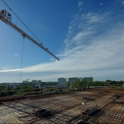 Biuro Inżynierskie Jakub Szewczyk - Widok z góry na zbrojenie stropu w trakcie budowy, z żurawiem wieżowym w tle i panoramą miasta w oddali, słoneczny dzień.