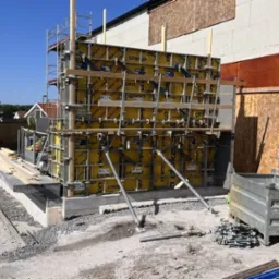 Szare betonowe fundamenty budynku w trakcie wylewania, z żółtymi szalunkami Doka i rusztowaniami, widoczne deski i elementy konstrukcyjne.