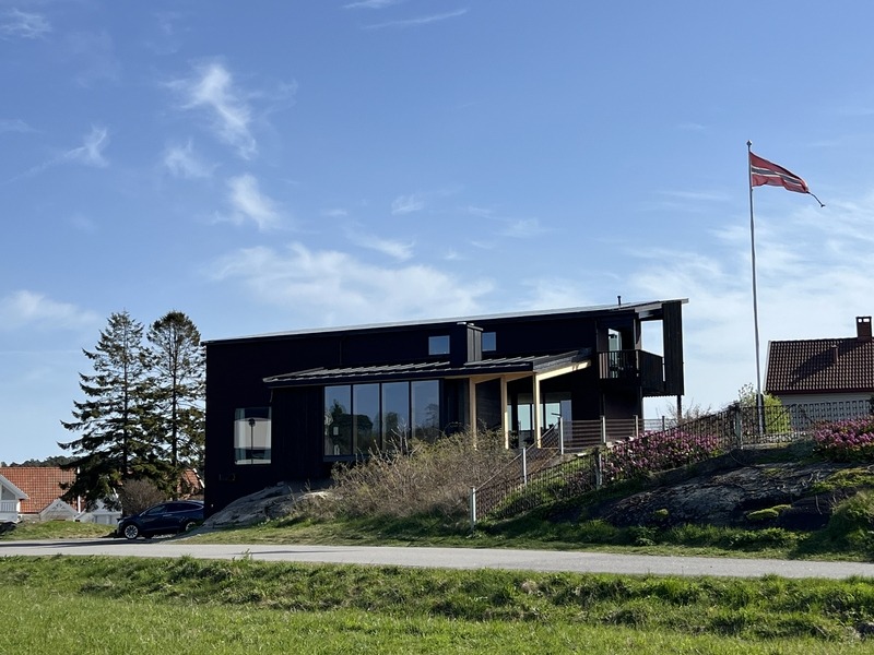 Nowoczesny dom z czarną elewacją, dużymi oknami i balkonem, otoczony zielenią i norweską flagą na maszcie.