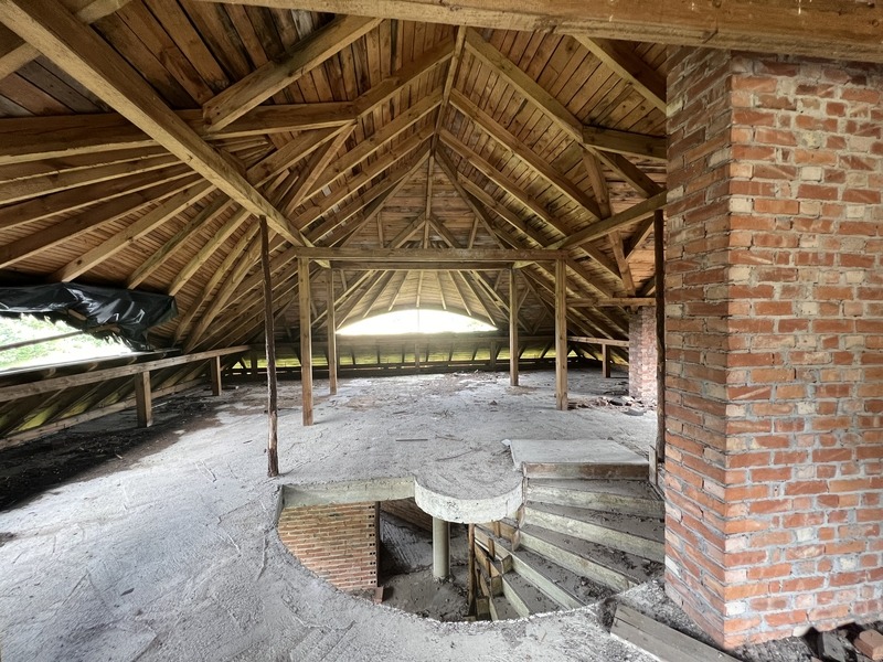 Wnętrze budynku w stanie surowym otwartym z drewnianą konstrukcją dachu, ceglanym kominem i betonową podłogą ze schodami spiralnymi prowadzącymi w dół.