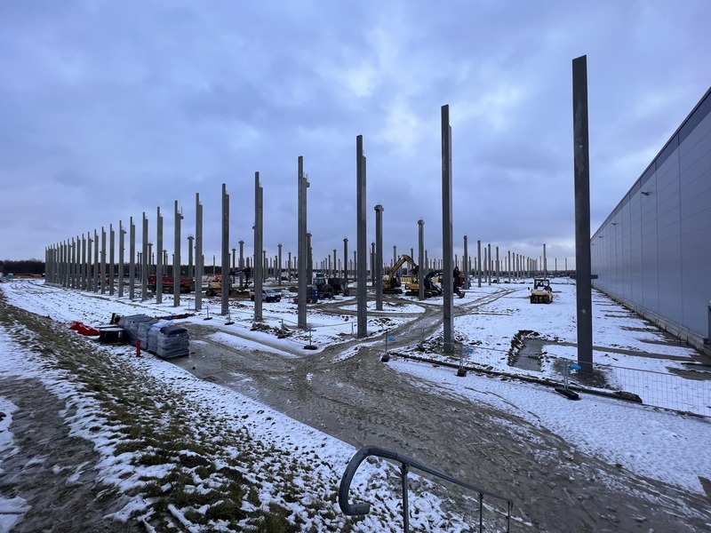 Wznoszenie konstrukcji hali przemysłowej: liczne betonowe słupy na zasypanym śniegiem placu budowy, widoczne maszyny budowlane w tle.