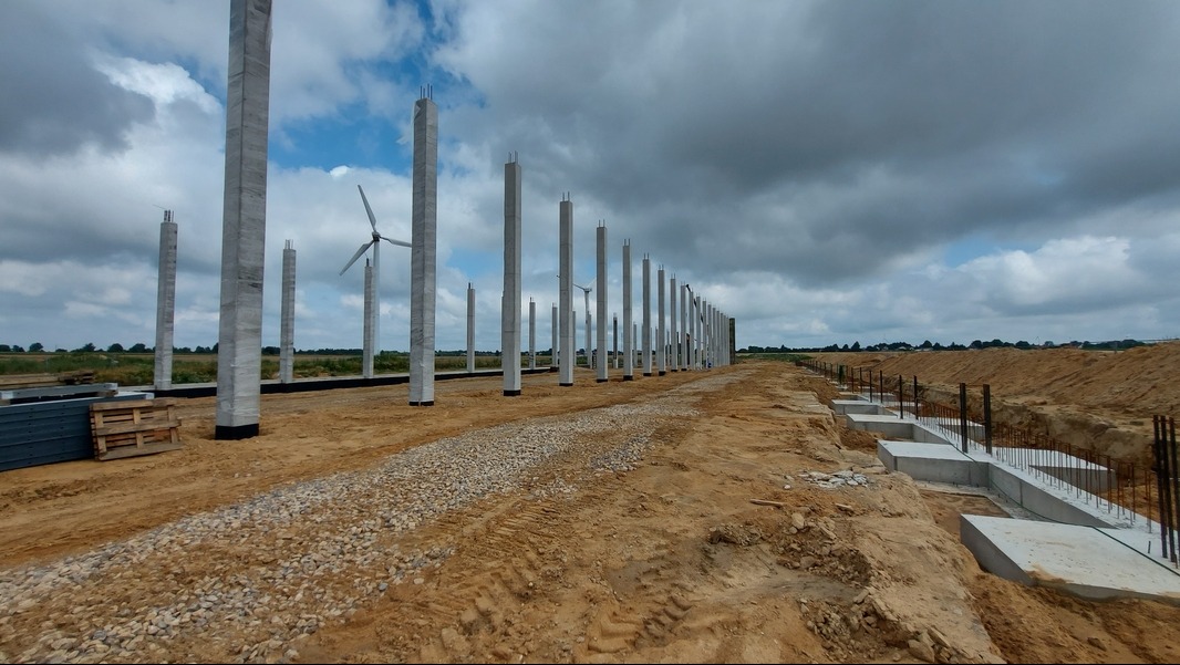 Wznoszenie konstrukcji szkieletowej hali przemysłowej: betonowe słupy, zbrojenia i fundamenty w wykopie, turbina wiatrowa w tle.