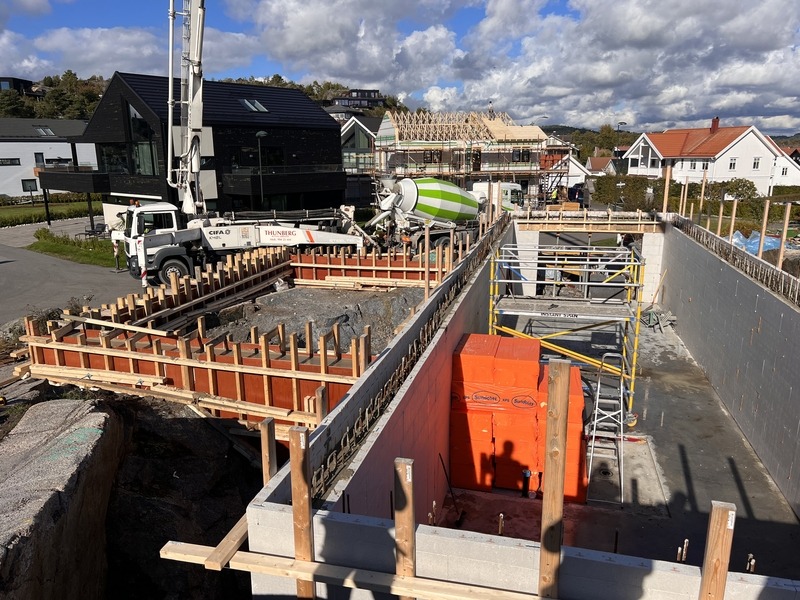 Fundamenty domu w trakcie budowy: betonowe ściany szalunkowe, zbrojenie, pompa do betonu Cifa i betoniarka na tle domów jednorodzinnych i pochmurnego nieba.