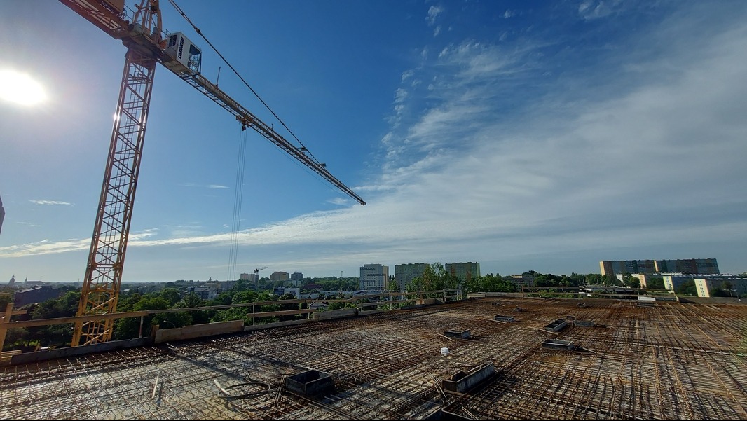 Widok z góry na zbrojenie stropu w trakcie budowy, z żurawiem wieżowym w tle i panoramą miasta w oddali, słoneczny dzień.