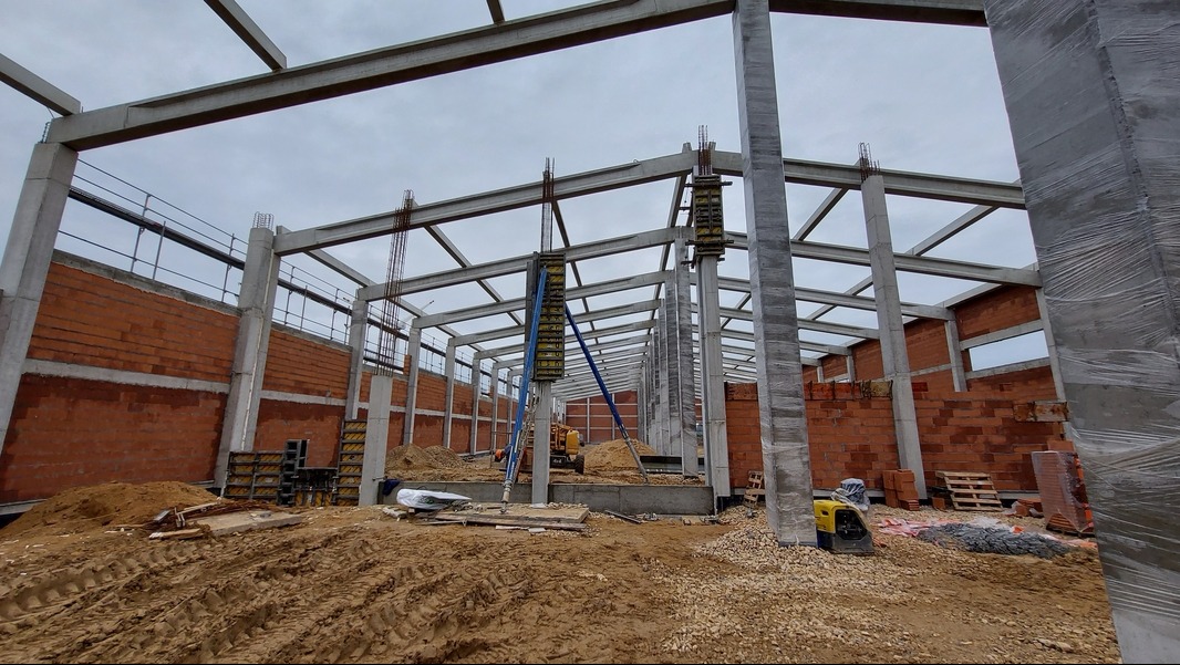 Wznoszenie konstrukcji hali z betonu i cegły, widoczne szalunki na słupach, plac budowy z gruzem i ziemią.