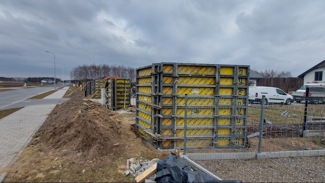 Szare, metalowe szalunki wypełnione żółtą okładziną, ustawione wzdłuż ogrodzenia posesji na etapie budowy fundamentów, widoczne samochody dostawcze i domy w tle.