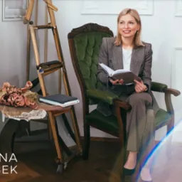 Kobieta w tweedowej marynarce siedzi w fotelu, trzyma otwarty notatnik i uśmiecha się, w tle sztaluga i stolik z dekoracjami.