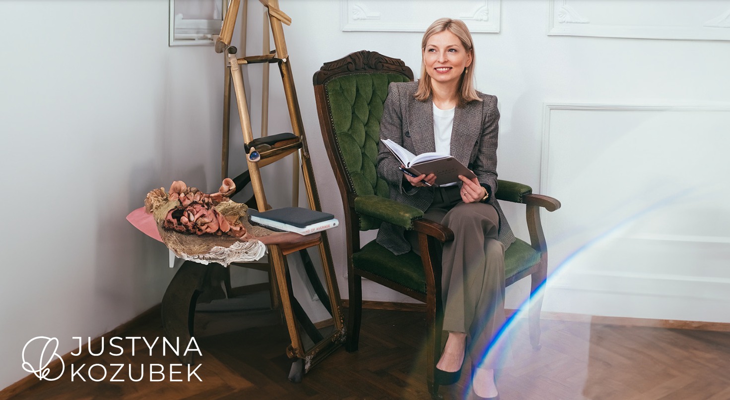 Kobieta w tweedowej marynarce siedzi w fotelu, trzyma otwarty notatnik i uśmiecha się, w tle sztaluga i stolik z dekoracjami.